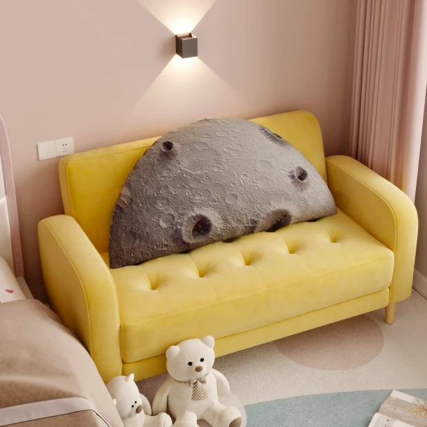 Ein großes, halbkreisförmiges Kissen in Grau mit einem 3D-Mondmotiv (Krater) liegt auf einem kleinen, gesteppten, gelben Sofa.