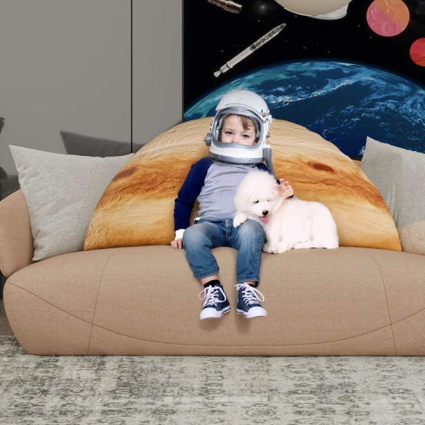 Ein entzückendes Foto von einem Kind mit Astronautenhelm und einem weißen Hund, der auf einem beigen Sofa mit einem riesigen 3D-Jupiter-Kissen sitzt.