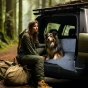 Eine Frau in Grün, mit Rucksack, sitzt im Wald neben dem offenen Kofferraum eines Geländewagens. Ein Hund sitzt neben ihr auf einer blau-weiß gestreiften Matte.