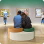 Ein Pärchen sitzt eng aneinander gekuschelt auf einem geometrischen modularen Pouf (in Grün, Orange und Beige) mitten in einer Kunstgalerie/einem Museum.