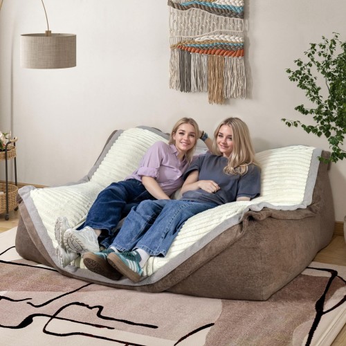 Zwei lächelnde Frauen entspannen sich auf einem großen, braun-beigen modularen Lounge-Sofa mit weichem, geripptem Stoff.