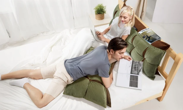Ein Paar liegt auf einem Bett und benutzt die grünen und orangen Kopfstützkissen mit Stauraum.