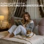 Frau sitzt mit Hund auf dem Bett und lehnt sich an ein rosafarbenes Vintage-Samtkissen.