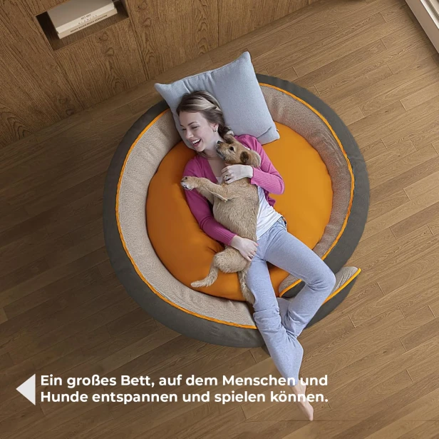 Eine Frau, die einen kleinen Hund in einem sehr großen, runden, grau-orangenen Hundebett von oben streichelt.