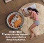 Der Hund schläft zusammengerollt in einem warmen, runden Bett in Grau und Orange, während die Frau neben ihm auf dem Teppich schläft.