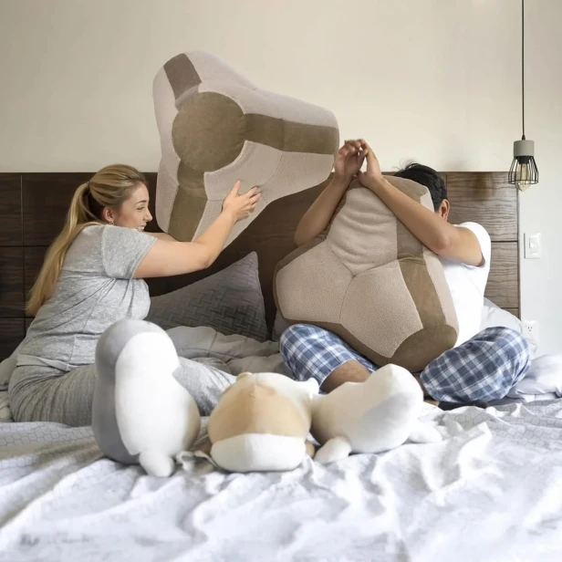 Ein paar Erwachsene veranstalten im Bett eine lustige Kissenschlacht mit großen, geometrischen Kissen.